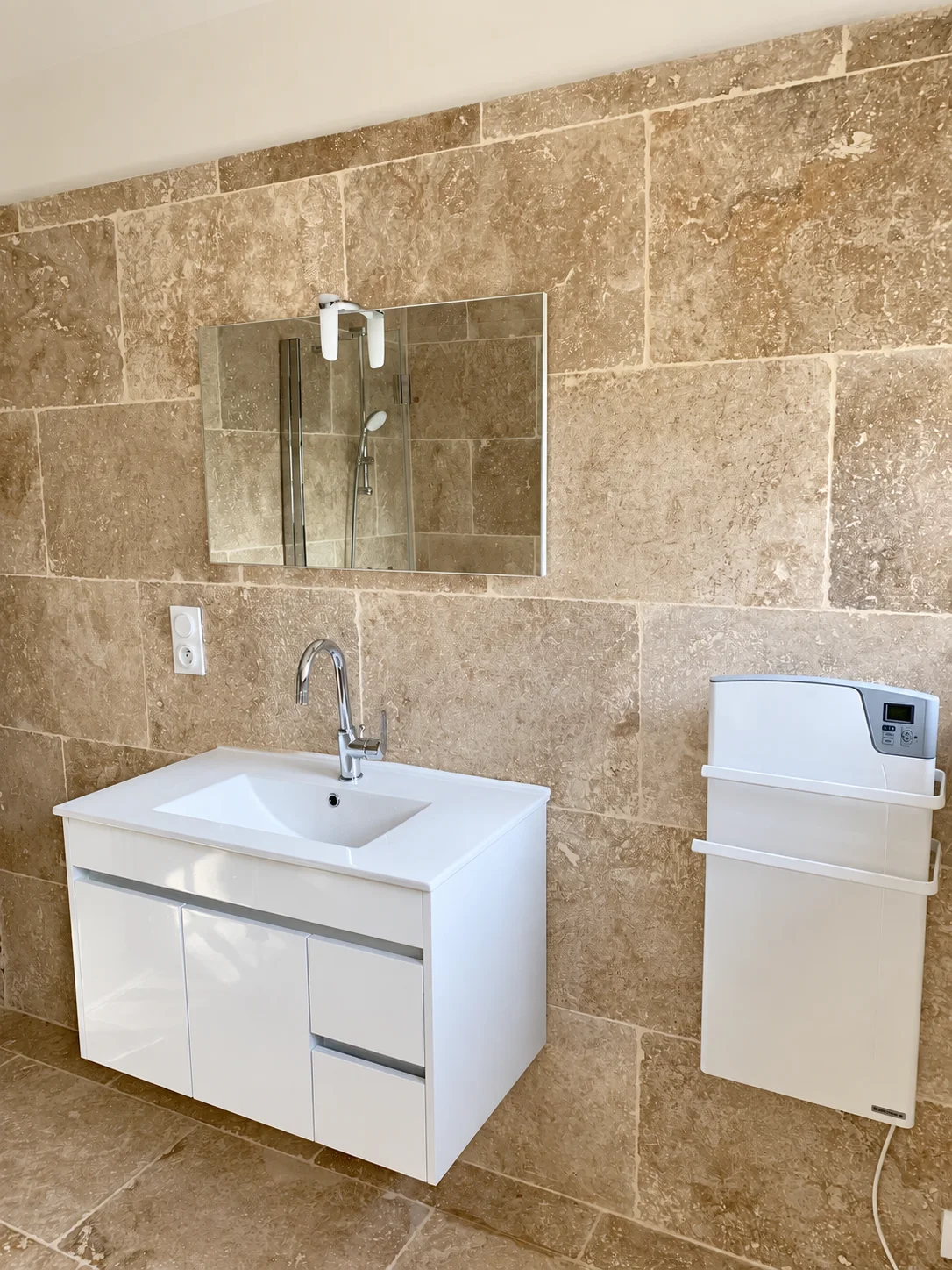 Meuble vasque blanc suspendu, miroir et sèche-serviettes mural sur carrelage beige.